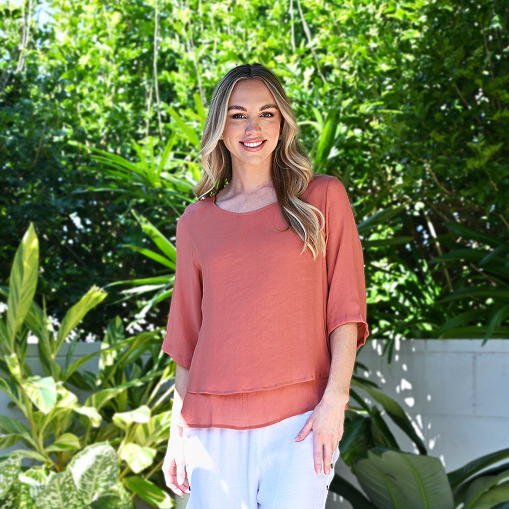 Woman wearing a coral top and white pants standing in front of green foliage
